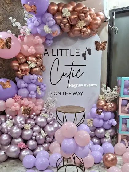 A beautiful baby shower decoration in pastel pink, purple, and chrome colors. The setup includes a custom backdrop, butterfly accents, and transparent 'BABY' boxes filled with balloons.