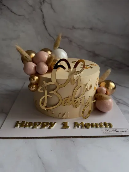 "Oh Baby!" This cake celebrates a baby's one-month milestone with a chic design, featuring a gold acrylic topper, and a cascade of pink and gold spheres.