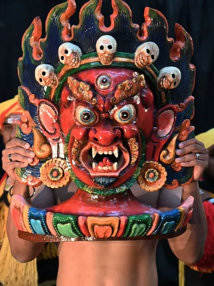 A fierce red mask from Nepal, held up during a performance. These hand-painted masks are designed to be vibrant and expressive, serving as powerful evil eye warders for your home's entrance.