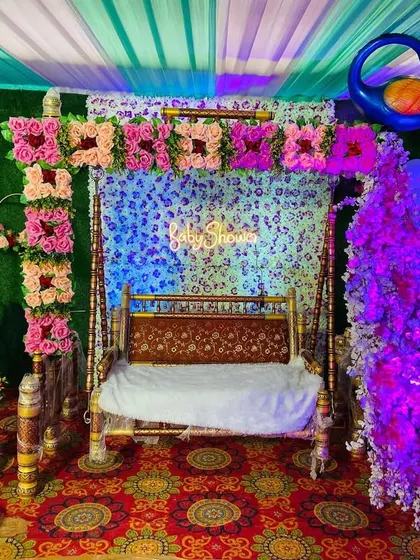 A close-up of a baby shower swing (jhula) decorated with pink flowers and set against a green backdrop with a neon sign.