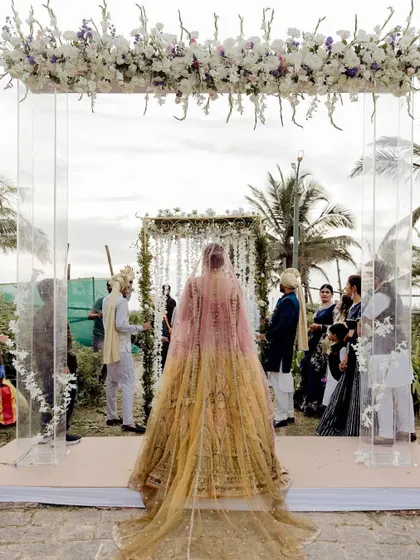 A bride makes her entrance towards a simple yet elegant mandap with hanging white florals, perfect for a beach wedding.