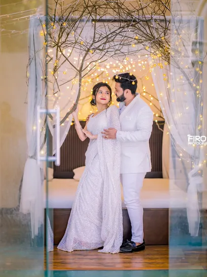 A stylish couple poses in all-white outfits against a backdrop of fairy lights and sheer curtains, creating a chic and modern pre-wedding portrait.