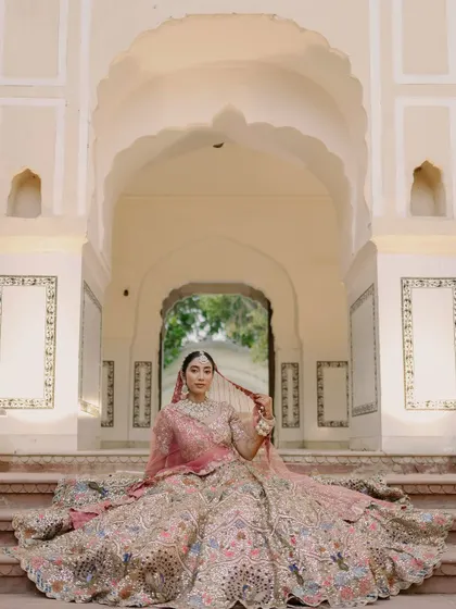 A regal bridal portrait showcasing the bride seated on the steps of a heritage property. Her magnificent, multi-colored lehenga is spread out, displaying its intricate details against the palace's classic arches, a perfect example of timeless wedding photography.