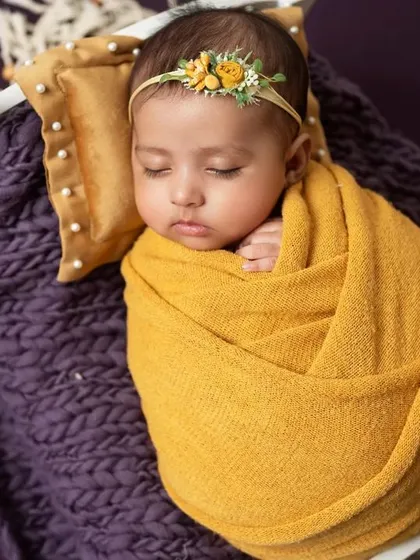 A swaddled newborn in a rose-like wrap, sleeping on a tiny bed with yellow flowers.