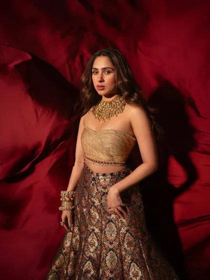 Pashmina Roshan looking like royalty against a rich red backdrop, wearing a magnificent gold choker and stacked kadas.