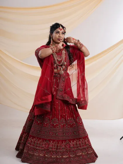 A stunning red bridal lehenga with intricate embroidery. The model is adorned with heavy traditional jewelry, showcasing a complete rental look for a bride.