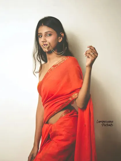 A confident and direct portrait of a model in a red saree and traditional nath, looking straight at the camera.
