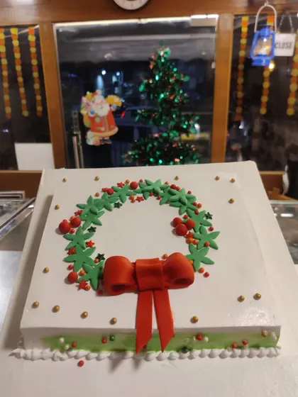 Our Christmas wreath cake is an elegant choice for your holiday table. The simple square cake is topped with a beautifully piped green wreath, red berry details, and a festive red bow.