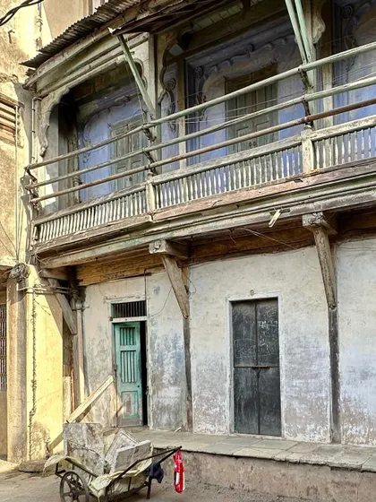 A facade at Majumdar Wada showing a wooden balcony and doorways. The varying colors and states of repair reflect the building's continuous, layered history of occupation and adaptation.