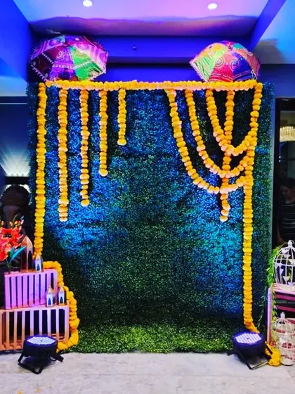 A simple yet elegant photo booth setup for a Mehndi or Haldi function. An artificial green grass wall is draped with strings of yellow marigolds and topped with two colorful umbrellas, creating a fresh and festive look.