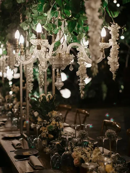 A close-up of the tablescape from a beautiful wedding dinner. The setting features elegant chandeliers dripping with white wisteria, creating a magical, intimate atmosphere for guests.