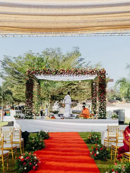 An open-air mandap that embraces the natural beauty of its surroundings. Set against a backdrop of hills, this structure, adorned with red and pink roses, offers a grand yet organic setting for a daytime wedding ceremony in Tumkur.