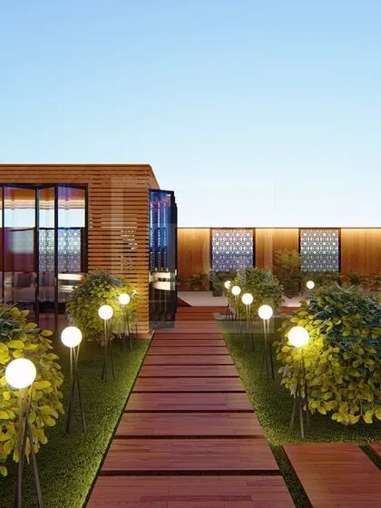 A pathway in the Gurgaon project, leading towards a glass-enclosed seating area, surrounded by lush bushes and globe lights.