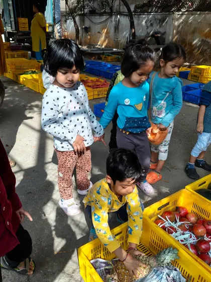 Children carefully inspect pineapples and apples in their crates. These trips help build vocabulary and a connection to healthy, fresh food.