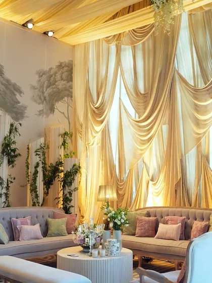 A luxurious lounge area set against a backdrop of hand-painted wallpaper and dramatic golden drapes. The pastel-colored sofas and floral arrangements create a soft, inviting space.