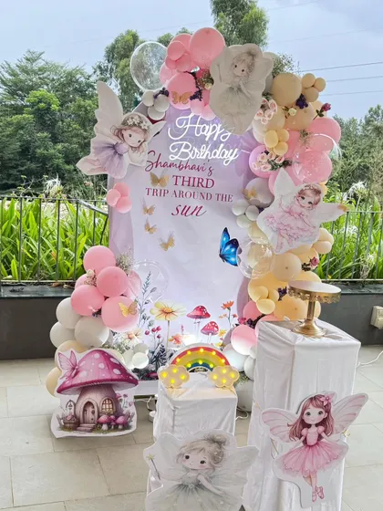 A whimsical fairy-themed party for a third birthday, set up on a beautiful balcony. This decor includes a pastel balloon garland, a custom backdrop, and adorable cutouts of fairies, a mushroom house, and a rainbow.
