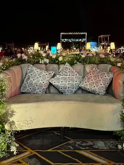 A romantic outdoor reception seating area. The sofa is surrounded by a lush arrangement of greenery and white roses, creating a beautiful focal point.