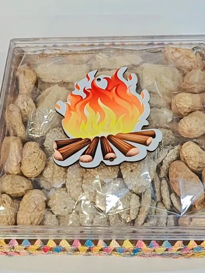 A simple and festive Lohri gift box with a clear lid and a bonfire sticker, filled with peanuts and sesame rewari.