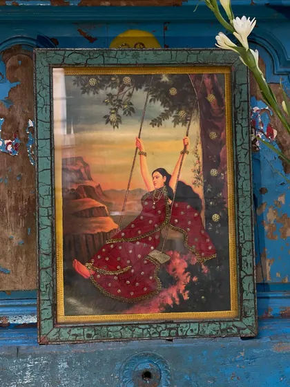 A cloth oleograph of a woman on a swing, set against a distressed blue door. The vintage print is enhanced with embroidery, creating a beautiful piece that mixes tradition with a fresh, textured look.
