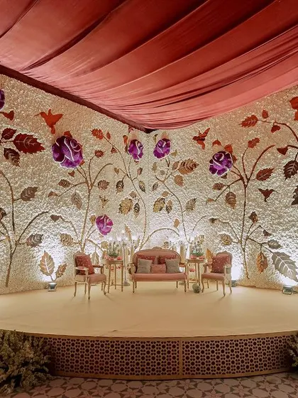 This magnificent stage backdrop is a custom-made flower wall, with a base of white carnations and a design of climbing roses made from purple and metallic-gold dyed petals.