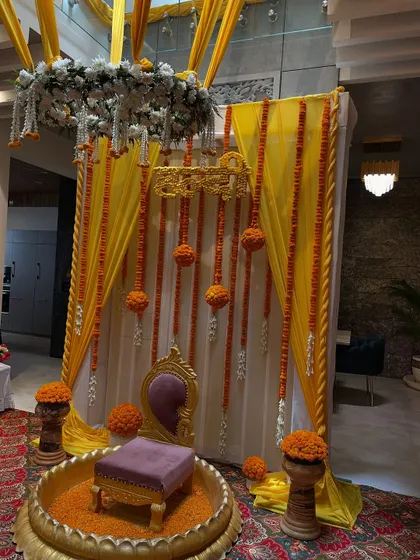 An elegant and traditional Haldi seating area. The setup features a beautiful backdrop with yellow drapes, marigold garlands, and a classic 'baithak' style seat for the ceremony.