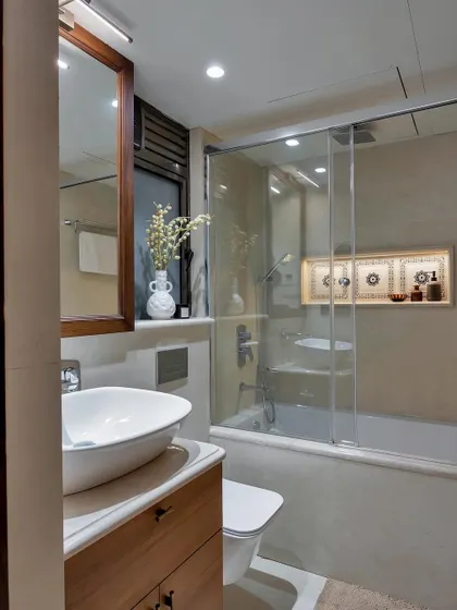 This elegant modern bathroom has a cozy ambiance. It features a sleek wooden vanity, a vessel sink, and a glass-enclosed bathtub with a built-in niche for toiletries, blending style and function perfectly.