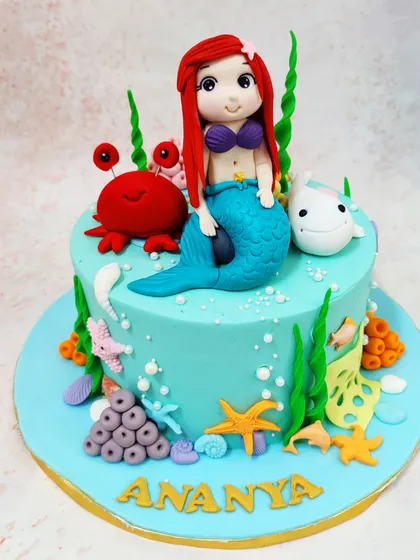 We dive under the sea with this enchanting Little Mermaid cake. A sweet Ariel figure sits beside her friend Flounder and a cheerful crab, surrounded by seaweed and starfish on a beautiful aqua-blue cake, creating a magical underwater scene.