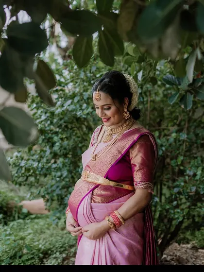 A candid and serene outdoor maternity portrait. The mother-to-be, dressed in a beautiful pink and purple saree, shares a quiet moment of reflection surrounded by the soft light and natural greenery.