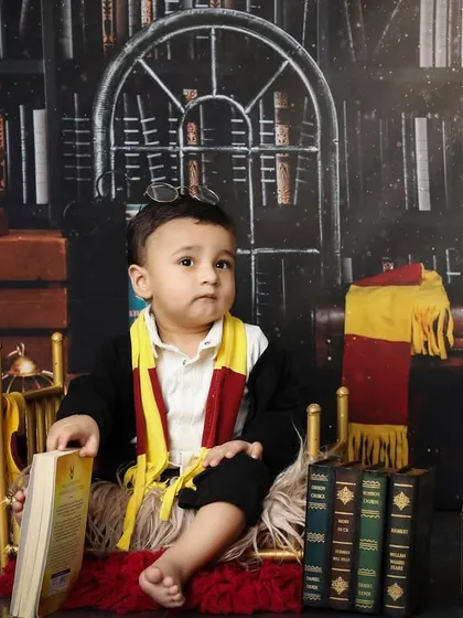A moment of quiet contemplation for our little wizard. This pose shows off the full Gryffindor-style costume against the rich, dark library backdrop, perfect for a themed baby portrait.
