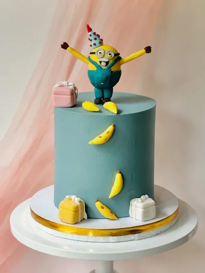 A happy Minion celebrating with gift boxes and bananas. The simple teal buttercream cake provides a great backdrop for the playful, hand-sculpted fondant character.