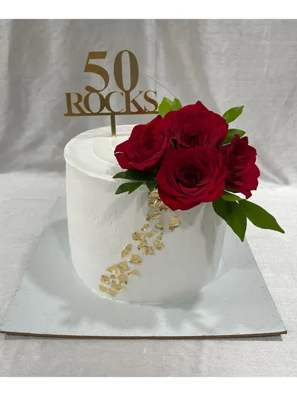 A classy cake for a 50th birthday with the message "50 Rocks". The clean white finish is accented with gold leaf and a bouquet of fresh red roses.