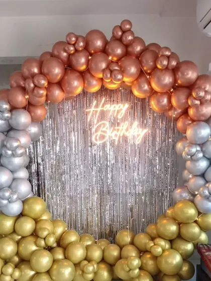 A glamorous circular balloon frame in rose gold, silver, and gold chrome balloons, set against a silver shimmer curtain with a neon sign.