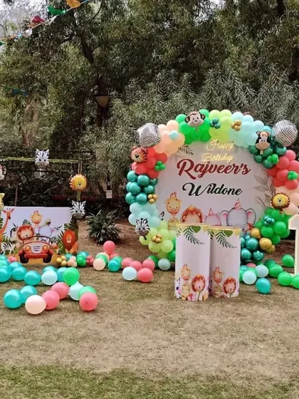 A "Wild One" jungle-themed first birthday party in a beautiful garden setting. The decoration includes a personalized backdrop, balloon garlands with animal characters, and themed props scattered across the lawn.