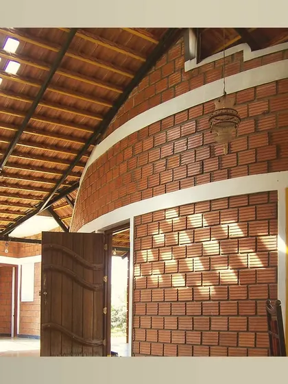 A curved wall made of hollow clay blocks creates a dynamic, flowing entrance. The exposed material, combined with a sloping roof and a handcrafted wooden door, celebrates the beauty of neo-traditional design.