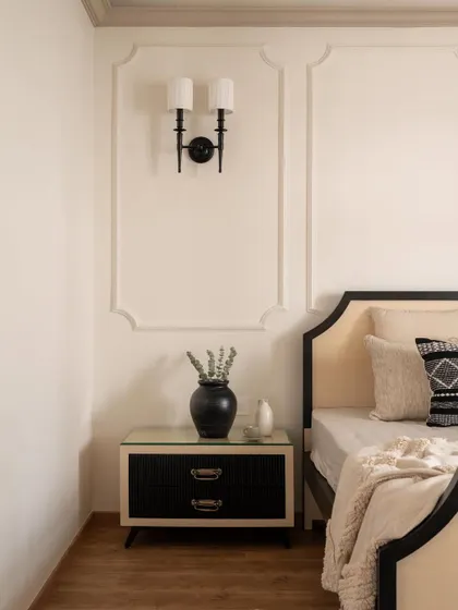 A classic black and cream bedroom from the Riviera Maison project. The bed features a gracefully curved headboard, flanked by a modern black nightstand and a traditional wall sconce.
