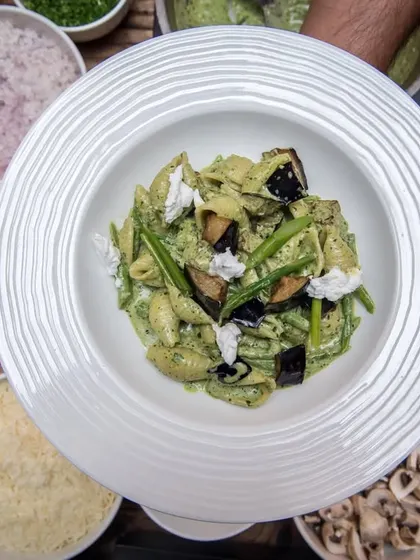 A hearty bowl of pesto pasta with roasted eggplant, surrounded by fresh preparation ingredients.