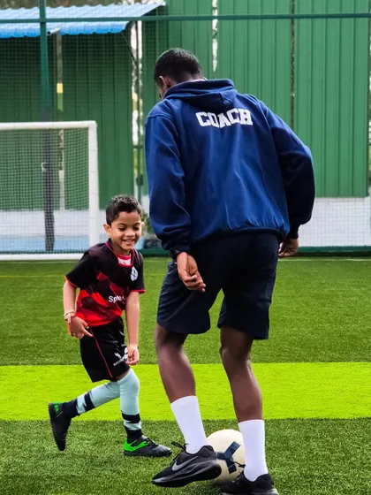A coach, clearly identified on his hoodie, engages one-on-one with a young player, demonstrating a move and making learning personal and fun.