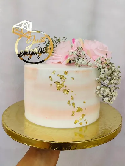 A delicate "Just Engaged" cake. The soft pink watercolor effect is accented with gold leaf, fresh pink roses, and a custom ring-shaped topper.