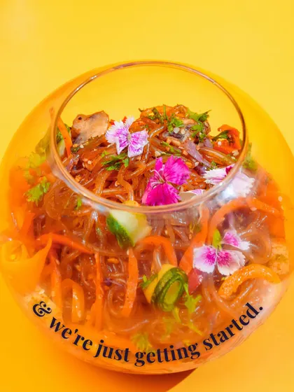 A close-up of my stunning glass noodle salad, filled with colorful vegetables and garnished with edible flowers. The text "and we're just getting started" hints at the endless creativity and flavor I bring to my vegetarian menu.