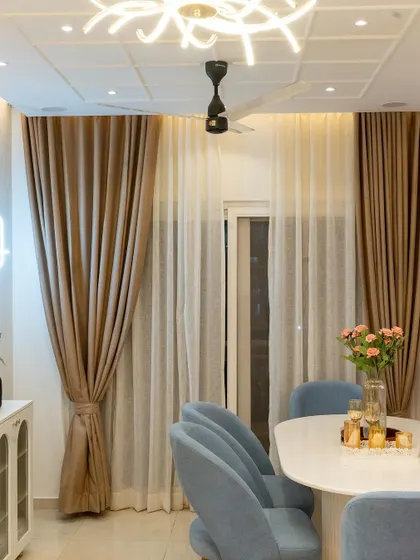 The dining area is simple yet elegant, with plush blue chairs, a white oval table, and beautiful floor-to-ceiling curtains that add a sense of luxury.