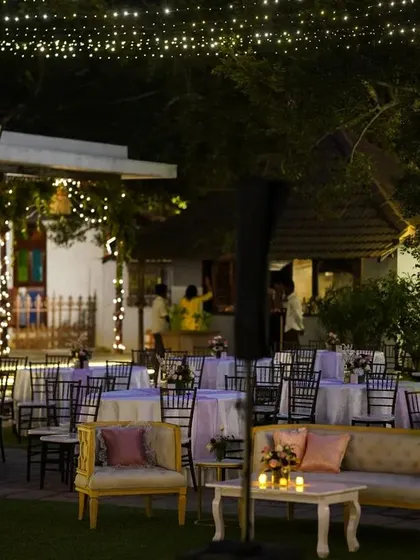 An enchanting garden dining setup. Tables are arranged under a canopy of trees adorned with hanging lanterns and fairy lights, creating a magical and intimate atmosphere for an outdoor reception.