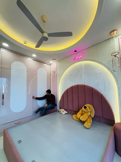A look at a dreamy kids' room in pink and white. The room features a bed with a large upholstered arch headboard, a neon name sign, and a sliding wardrobe with matching curved details.