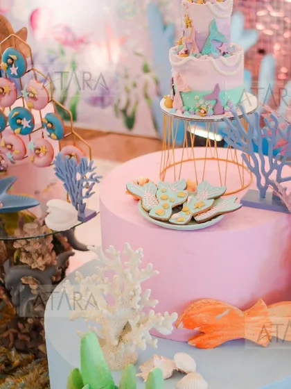 A detailed look at the dessert table from a mermaid-themed party. The setup includes a beautiful cake, themed cookies, and donuts, all displayed with coral and other sea-inspired props.