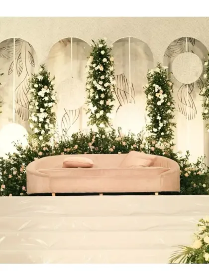 A clean and modern stage design using a white backdrop with minimalist line art. The decor is brought to life with vertical arrangements of white flowers and greenery, and a soft pink sofa adds a subtle touch of color.