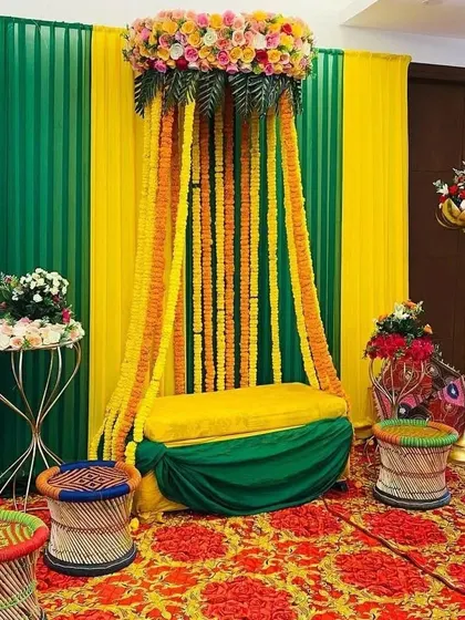 A traditional Mehendi seating area with a canopy of marigold strings. The setup includes colorful 'mooda' stools and a backdrop of green and yellow drapes.