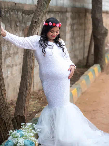 A lovely outdoor portrait of a mom-to-be in a white lace gown and floral crown, leaning against a tree.