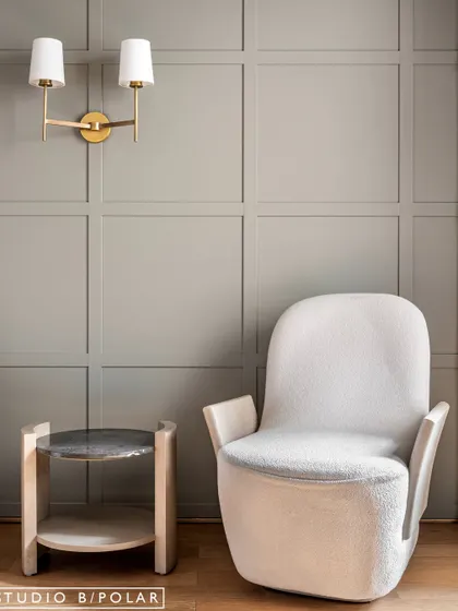 A cozy seating corner with a comfortable armchair and a small side table. The soft grey wall color and classic wall sconce create a serene and timeless feel.