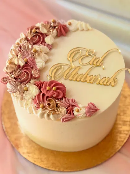 An elegant cake for Eid, featuring a crescent of intricately piped buttercream flowers in shades of dusty rose and cream, with a gold "Eid Mubarak" topper.