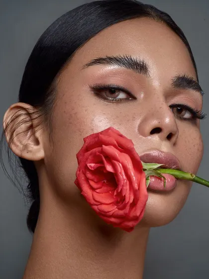 A romantic and modern beauty shot. The model holds a single red rose, its color providing a soft contrast to the minimal makeup and freckled skin. The pose is both strong and delicate.