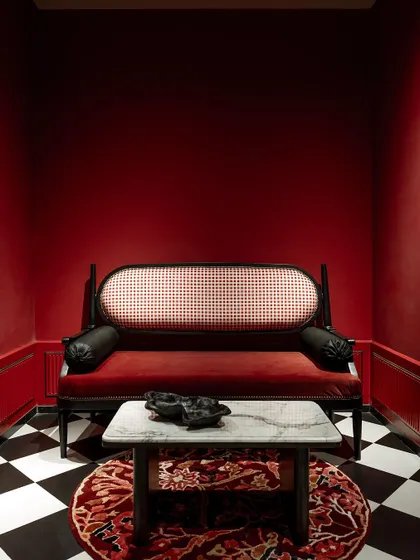 This vignette from our House of Goya experience center showcases our approach to maximalism. A bold, color-drenched red room creates a dramatic backdrop for a classic caned settee, a marble-topped table, and a patterned rug, demonstrating how curated layers can create a powerful mood.